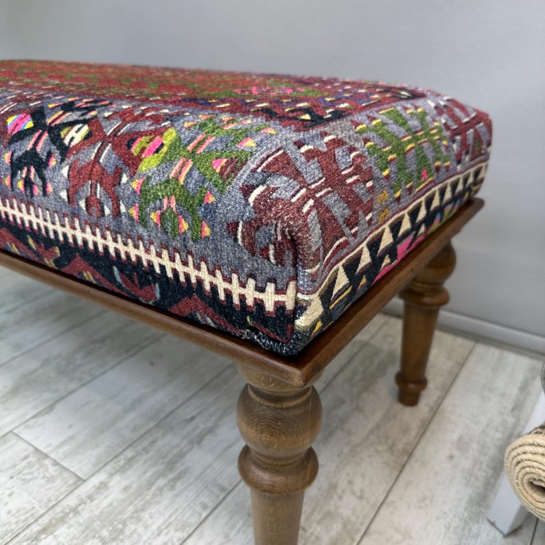 May include: A rectangular ottoman with a patterned textile top in shades of red, gray, and green, resting on four turned wooden legs. The textile features geometric and floral designs.