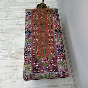 May include: A rectangular ottoman with a colorful, patterned textile top. The design features a central red field with green, tree-like motifs, and borders with geometric shapes in various colors. The ottoman is placed on a light-colored wooden floor.