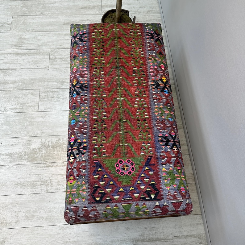 May include: A rectangular ottoman with a colorful, patterned textile top. The design features a central red field with green, tree-like motifs, and borders with geometric shapes in various colors. The ottoman is placed on a light-colored wooden floor.