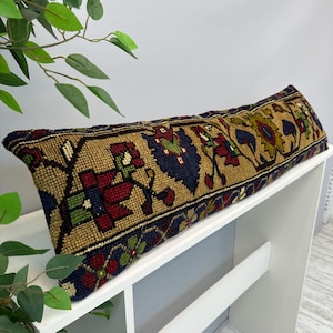 May include: A long rectangular lumbar pillow featuring a woven textile cover with a dense, multi-colored geometric and floral pattern in shades of mustard yellow, deep navy blue, dark red, and forest green on a textured, knotted surface.