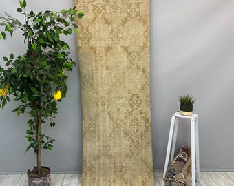 May include: A long, rectangular, beige rug with an intricate floral pattern. The rug is hanging against a gray wall. A potted lemon tree and a small white plant stand with rolled rugs are in the background.