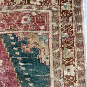 May include: Close-up of a section of a vintage rug with a distressed appearance. The rug features a central design in shades of red and teal, with intricate patterns and a border in brown and beige. The rug's texture is visible.