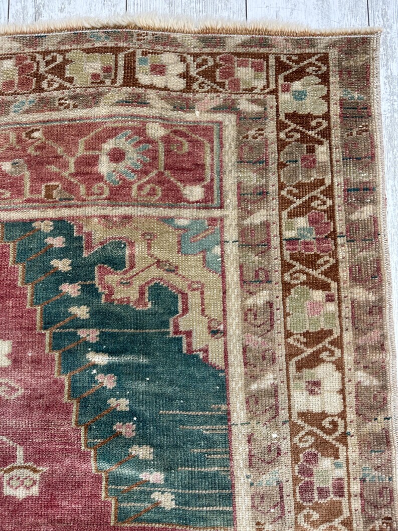 May include: Close-up of a section of a vintage rug with a distressed appearance. The rug features a central design in shades of red and teal, with intricate patterns and a border in brown and beige. The rug's texture is visible.