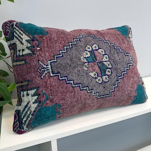 May include: A rectangular throw pillow featuring a textured, woven surface with a deep magenta base color. The design includes a large, central gray diamond shape with a jagged border, accented by teal and white geometric patterns at the corners.