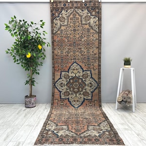 May include: A long, rectangular runner rug featuring a complex, symmetrical pattern in shades of terracotta, navy blue, and cream. The design includes a large central medallion and intricate geometric borders, with a fringed edge at the bottom.