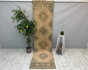 May include: A long, rectangular runner rug featuring a tan base color with a repeating pattern of dark brown, diamond-shaped medallions and intricate floral borders. The rug is displayed hanging vertically, showcasing its full length and design.