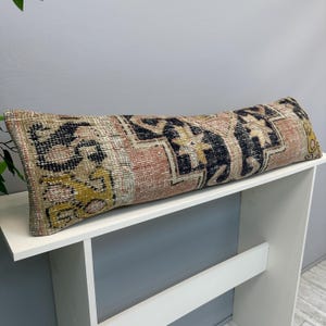 May include: A long, rectangular lumbar pillow featuring a woven textile cover with a distressed, multi-colored pattern in shades of faded pink, deep navy blue, mustard yellow, and beige, displaying intricate geometric and floral-inspired motifs.