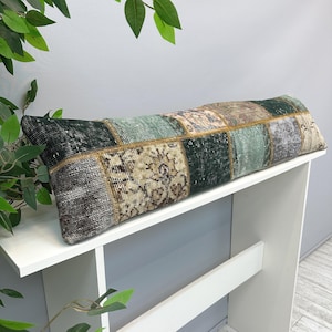 May include: A long, rectangular pillow with a patchwork design. The pillow features squares of various colors, including green, beige, and gray, with intricate patterns. The pillow is placed on a white surface, showcasing its decorative appeal.