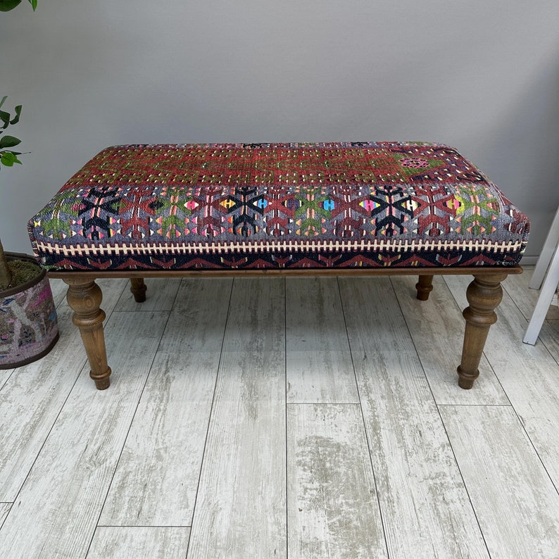 May include: A rectangular ottoman with a patterned textile top in shades of red, green, and blue. The wooden frame has turned legs. The textile features geometric designs and a dark border.