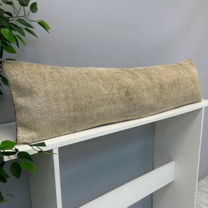 May include: A long, rectangular lumbar pillow featuring a textured, woven fabric in a neutral beige and tan color palette. The surface displays a subtle, faded pattern with visible cross-stitch details and a worn, vintage-inspired appearance.