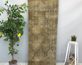 May include: A long, rectangular, distressed rug with a faded, ornate pattern in shades of brown and beige. The rug is hanging against a gray wall, showcasing its intricate design and aged appearance. The rug's dimensions are not visible.