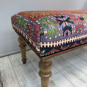 May include: Upholstered ottoman with a patterned textile top in shades of red, green, black, and gray. The wooden frame has turned legs. The ottoman is a decorative furniture piece.