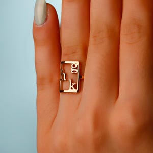 Może przedstawiać: Złoty pierścionek o prostokątnym kształcie, z literami "g" i "k" wewnątrz ramy. Pierścionek jest noszony na palcu, na jasnoniebieskim tle. Paznokcie są pomalowane na jasnozielony kolor.