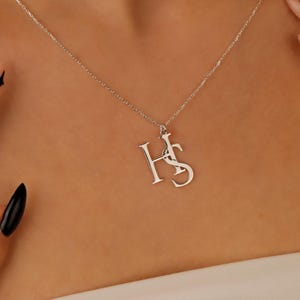 May include: A silver necklace with the initials "HS" as a pendant. The necklace is worn on a person's neck. The person has long, black nails and nails with a star design.