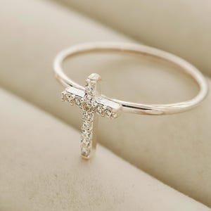 May include: A silver ring with a cross-shaped design. The cross is encrusted with small, clear gemstones. The ring is set against a neutral background with small, dried flowers.