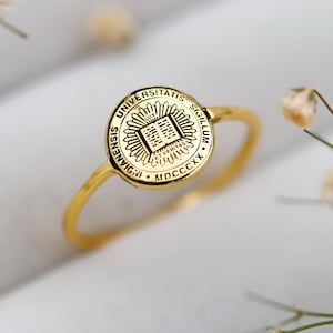 Może przedstawiać: Złoty pierścionek z okrągłym emblematem z napisem "UNIVERSITATIS SIGILLUM ENWIGII MDCCCXX". Pierścionek jest umieszczony na białym tle z delikatnymi białymi kwiatami.