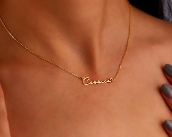 May include: A delicate gold necklace spelling out the name "Cassie" in cursive script. The necklace is worn around the neck, with the chain and pendant visible. The person's hands are near the necklace.