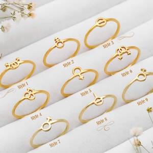 May include: Nine gold-colored rings with various symbol designs, including circles, lines, and stylized shapes. Each ring is labeled with a style number from 1 to 9. The rings are displayed on a white surface with small white flowers.
