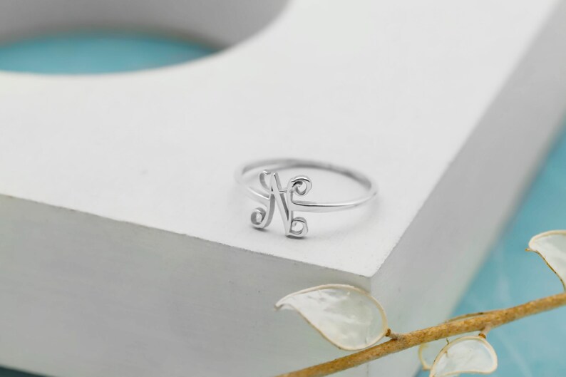 Puede incluir: Un anillo de tono plateado que presenta una letra N cursiva y estilizada como punto focal central. La banda es delgada y circular, conect&aacute;ndose con el dise&ntilde;o decorativo de la letra. El metal tiene un acabado liso y pulido que refleja la luz.