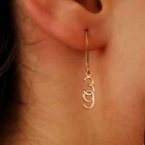 May include: Gold-toned dangle earrings with a number '3' charm. The earrings are shown on a person's ear.