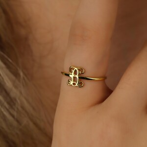 Puede incluir: Un anillo de oro con una banda fina y pulida y una letra B de monograma ornamentada en el centro. La letra est&aacute; dise&ntilde;ada con intrincados remolinos de caligraf&iacute;a que se conectan a la banda, creando un acabado met&aacute;lico dorado delicado.