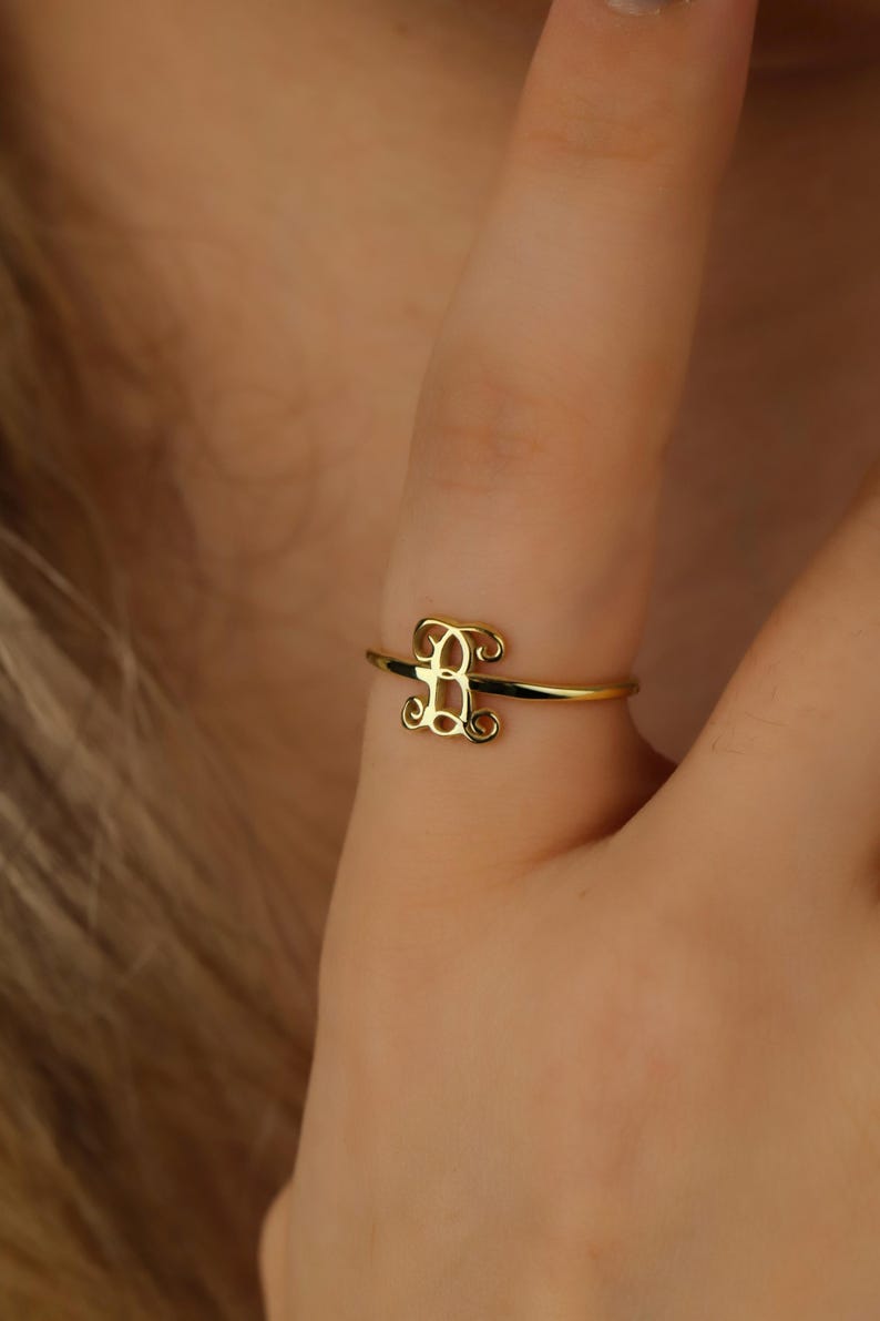 Puede incluir: Un anillo de oro con una banda fina y pulida y una letra B de monograma ornamentada en el centro. La letra est&aacute; dise&ntilde;ada con intrincados remolinos de caligraf&iacute;a que se conectan a la banda, creando un acabado met&aacute;lico dorado delicado.