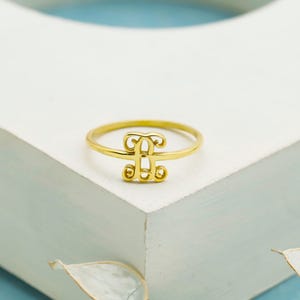 Puede incluir: Un anillo de tono dorado que presenta un elemento de monograma central con un dise&ntilde;o de letra estilizada. La banda es delgada y circular, sosteniendo la pieza central decorativa inicial que est&aacute; elaborada con l&iacute;neas curvas y un acabado pulido.