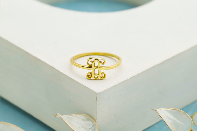 Puede incluir: Un anillo de tono dorado que presenta un elemento de monograma central con un dise&ntilde;o de letra estilizada. La banda es delgada y circular, sosteniendo la pieza central decorativa inicial que est&aacute; elaborada con l&iacute;neas curvas y un acabado pulido.