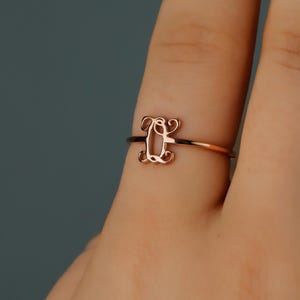 Puede incluir: Un anillo de oro rosa con una banda fina y pulida y un elemento decorativo central con forma de letra Q en una caligraf&iacute;a cursiva estilizada. El metal tiene un brillo met&aacute;lico c&aacute;lido y un acabado de superficie liso y reflectante.