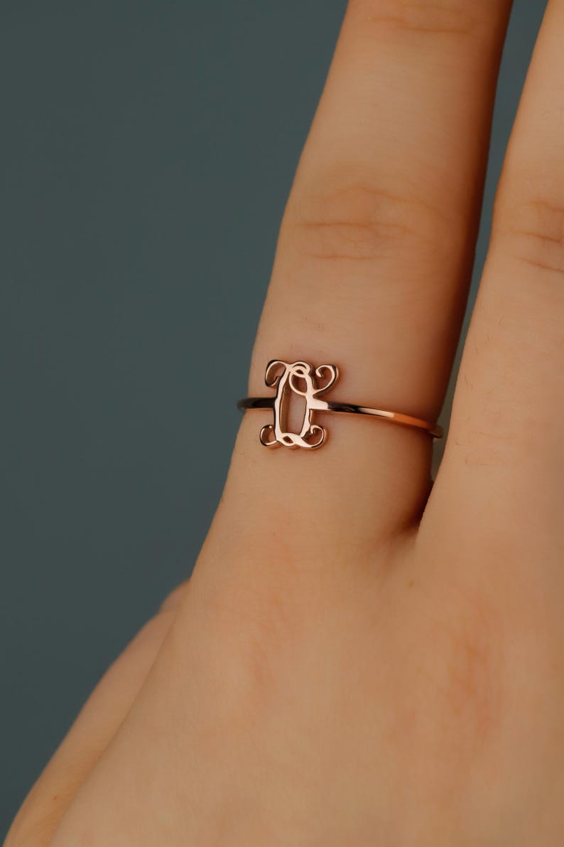 Puede incluir: Un anillo de oro rosa con una banda fina y pulida y un elemento decorativo central con forma de letra Q en una caligraf&iacute;a cursiva estilizada. El metal tiene un brillo met&aacute;lico c&aacute;lido y un acabado de superficie liso y reflectante.
