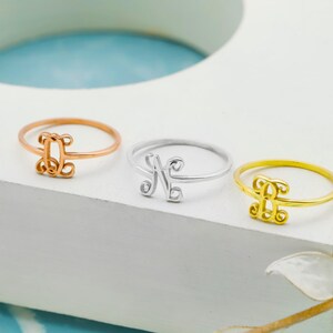 Puede incluir: Tres anillos finos de metal con una letra monograma decorativa en cursiva. El conjunto incluye un anillo de oro rosa con la letra J, un anillo de plata con la letra N y un anillo de oro con la letra J, cada uno con una banda circular.