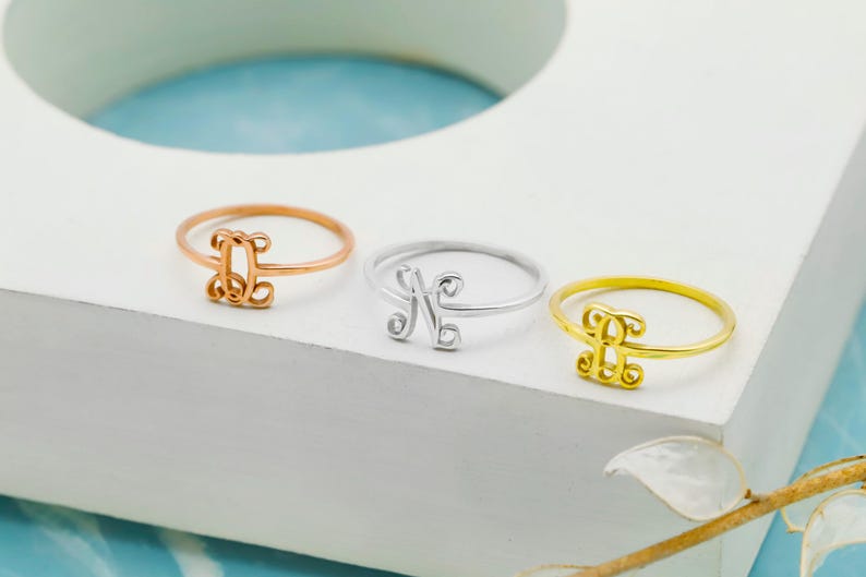 Puede incluir: Tres anillos finos de metal con una letra monograma decorativa en cursiva. El conjunto incluye un anillo de oro rosa con la letra J, un anillo de plata con la letra N y un anillo de oro con la letra J, cada uno con una banda circular.