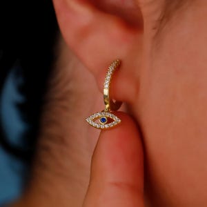 May include: A gold-colored hoop earring with a dangling eye charm. The hoop is encrusted with small, clear crystals. The eye charm features a blue center and is outlined with clear crystals, creating a sparkling effect. Jewelry.