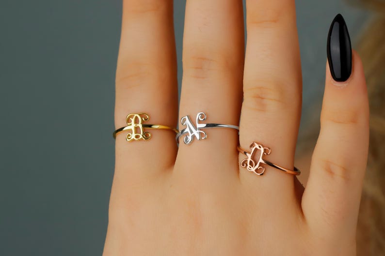 Puede incluir: Tres anillos finos de metal en los dedos, cada uno con una letra decorativa en cursiva. Un anillo es dorado con la letra B, otro es plateado con la letra N y el tercero es de oro rosa con la letra Q, todos con un acabado pulido.