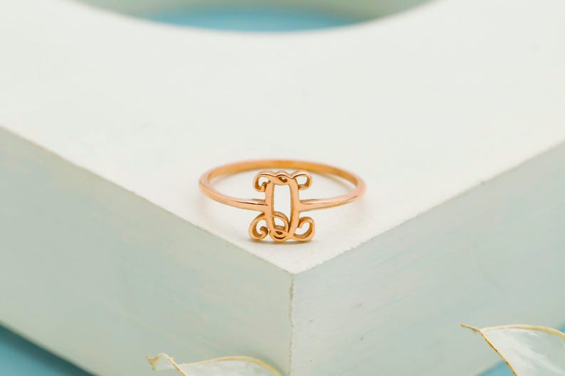 Puede incluir: Un anillo de oro rosa que presenta un elemento decorativo central con forma de letra D, dise&ntilde;ado con un intrincado trabajo de metal en estilo filigrana. La banda es delgada y circular, manteniendo un acabado met&aacute;lico uniforme en toda la pieza.