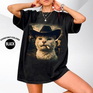 May include: A black short-sleeved t-shirt featuring a graphic print of a cat wearing a cowboy hat with a cigarette in its mouth, set against a farm background with cows. The text Comfort Colors Black is printed on the left side of the shirt.
