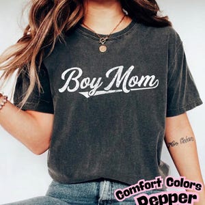 Peut inclure: Un t-shirt à manches courtes gris anthracite avec une impression graphique blanche vieillie indiquant Boy Mom dans une police cursive. Le vêtement présente un col rond et une finition délavée, porté avec un jean en denim bleu.