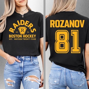 May include: Black t-shirt featuring gold text: "RAIDERS BOSTON HOCKEY". The back of the shirt shows "ROZANOV 81" in gold. The shirt is paired with blue jeans.