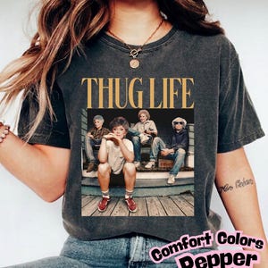 May include: A charcoal grey short-sleeve t-shirt featuring a graphic print of four individuals sitting on wooden steps. Above the image, the text THUG LIFE is printed in a bold, gold-coloured serif font. The shirt has a faded, vintage-style texture.