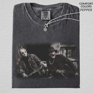 May include: A charcoal gray crew neck t-shirt featuring a graphic print of two horror movie characters, one wearing a hockey mask and holding a blade, the other in a fedora with clawed hands. The shirt includes a silver chain necklace.