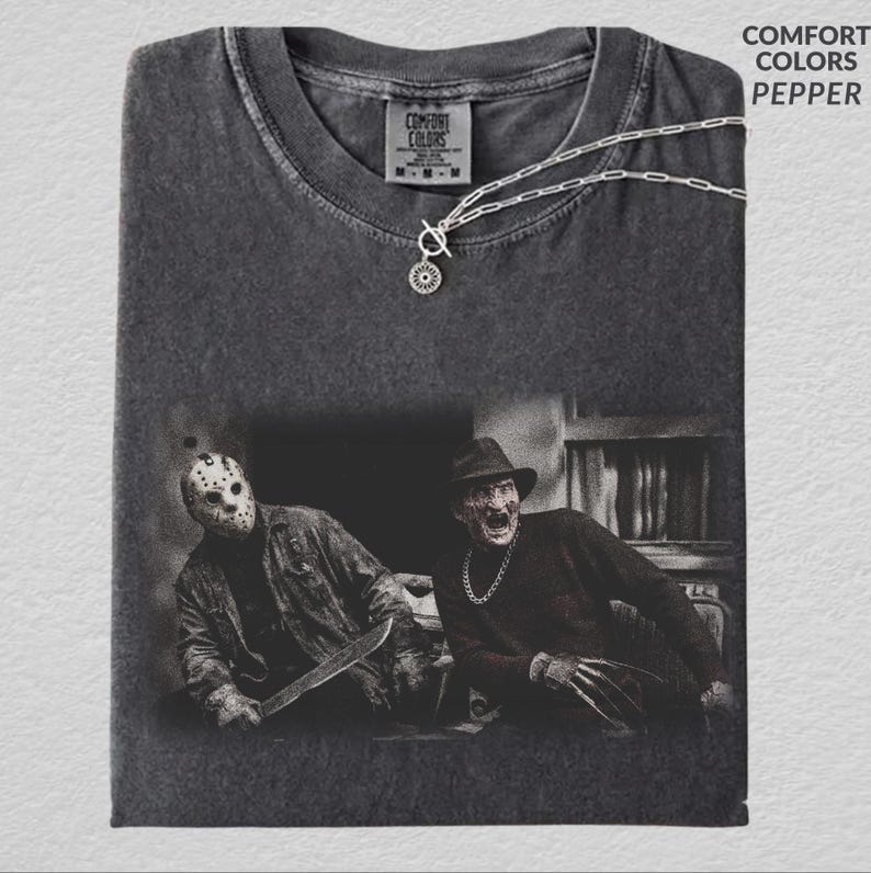 May include: A charcoal gray crew neck t-shirt featuring a graphic print of two horror movie characters, one wearing a hockey mask and holding a blade, the other in a fedora with clawed hands. The shirt includes a silver chain necklace.