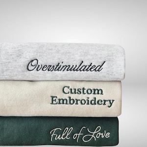 May include: A stack of three folded sweatshirts. The top is light grey with black embroidered text reading Overstimulated. The middle is cream with dark green embroidered text reading Custom Embroidery. The bottom is dark green with white text.