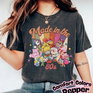 May include: A charcoal grey short-sleeve t-shirt featuring a graphic print of various 1980s cartoon characters. The design includes the text "Made in the 80s" above a colourful rainbow background with stars and multiple animated figures.