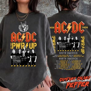 May include: A charcoal grey t-shirt featuring a graphic print of the band AC/DC. The front displays the band logo, the text PWR UP, and a black and white photo of the band members. The back lists the 2026 tour dates for South and North America.