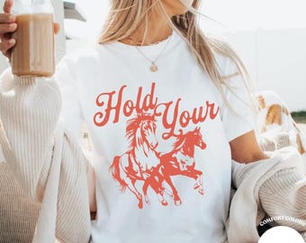May include: A white short-sleeve t-shirt featuring a red graphic print of two running horses and the text "Hold Your" in a stylized font. The shirt is layered under a beige cardigan and includes a small circular label reading "Comfort Colors White".