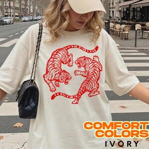 May include: An ivory short-sleeved t-shirt featuring a large red graphic of two tigers facing each other in a circular formation. The shirt is paired with a black quilted shoulder bag with a gold chain strap and a beige baseball cap.