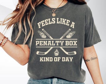 May include: Dark grey t-shirt with the text "Feels Like A Penalty Box Kind of Day" in a distressed font. The design includes crossed hockey sticks and a hockey puck. The shirt is a casual wear item.