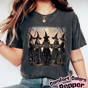 Peut inclure: Un t-shirt gris anthracite à manches courtes avec une impression graphique représentant cinq silhouettes portant des chapeaux pointus et de longues robes debout en cercle. Le texte "Girls Will Be Girls." est imprimé au centre du motif.
