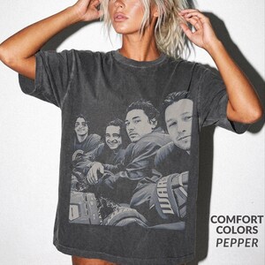 Peut inclure: Un t-shirt gris anthracite à manches courtes avec une impression graphique vintage en noir et blanc représentant quatre personnes. Le vêtement présente un aspect délavé et usé, avec le texte COMFORT COLORS PEPPER imprimé dans le coin.