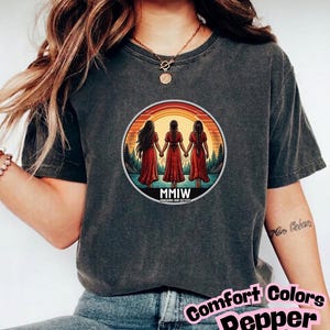 May include: Dark grey t-shirt with a circular graphic of three figures in red dresses holding hands, set against a sunset and forest backdrop. The text "MMIW" is below the figures. The shirt also has the text "Comfort Colors Pepper".